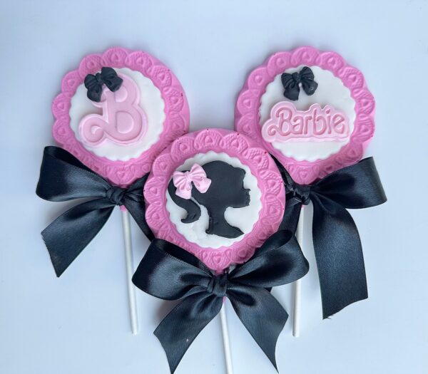 A set of three decorative lollipops in pink and white with black ribbons, featuring a silhouette, a "B" letter, and "Barbie," from the 12-Barbi Chocolate Lollipops collection for parties or favors.