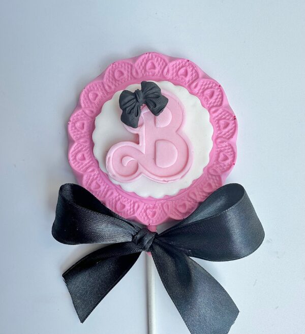 A pink and white Barbi chocolate lollipop with a letter "B," adorned with a black bow and ribbon, perfect for birthday parties or as party favors.