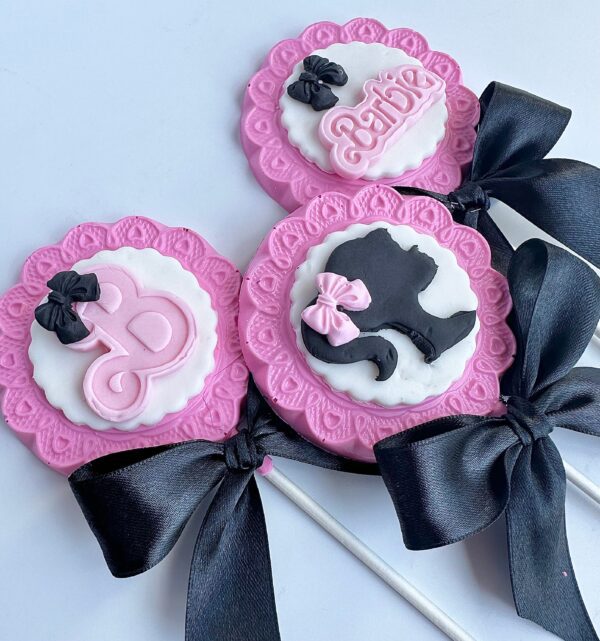 A set of three pink and white chocolate lollipop cookies with black bow decorations, featuring silhouette designs and the word "Barbie" from the 12-Barbi Chocolate Lollipops collection, perfect for parties.