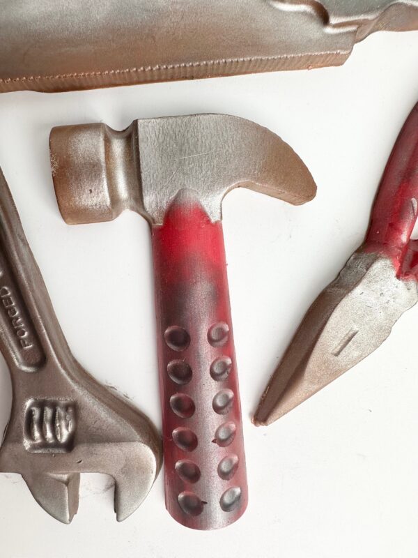 A close-up of a chocolate tool set features a red and grey hammer, adjustable wrench, and pliers on a white surface. This 5 Piece Chocolate Tool Gift Set comes in a gold gift box perfect for Dad's birthday or any special occasion.