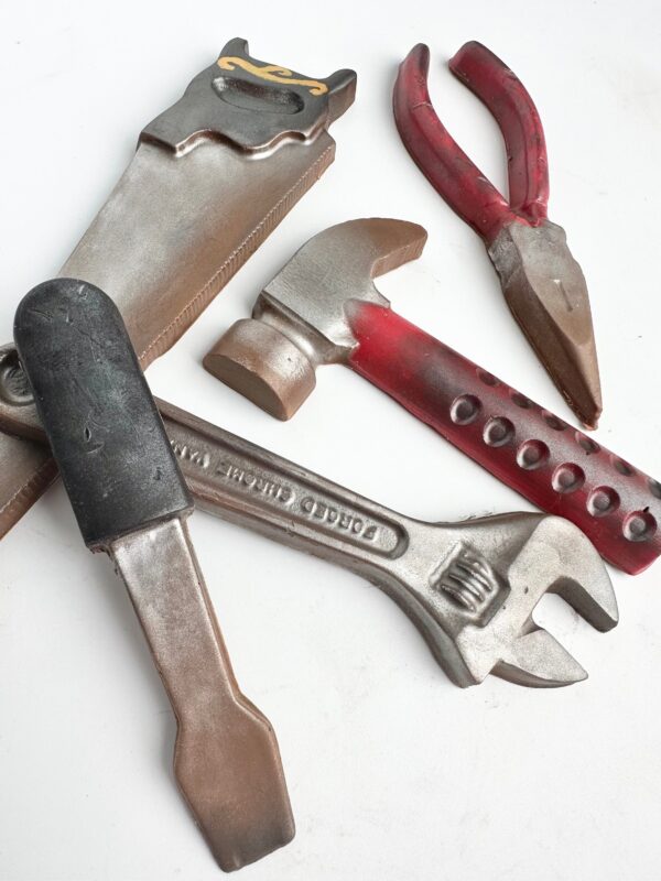 The 5 Piece Chocolate Tool Gift Set features wrench, hammer, saw, pliers, and utility knife shaped chocolates with cocoa powder dusting for realism on a white background, packaged in a gold gift box. Ideal for dad's birthday or any special occasion.