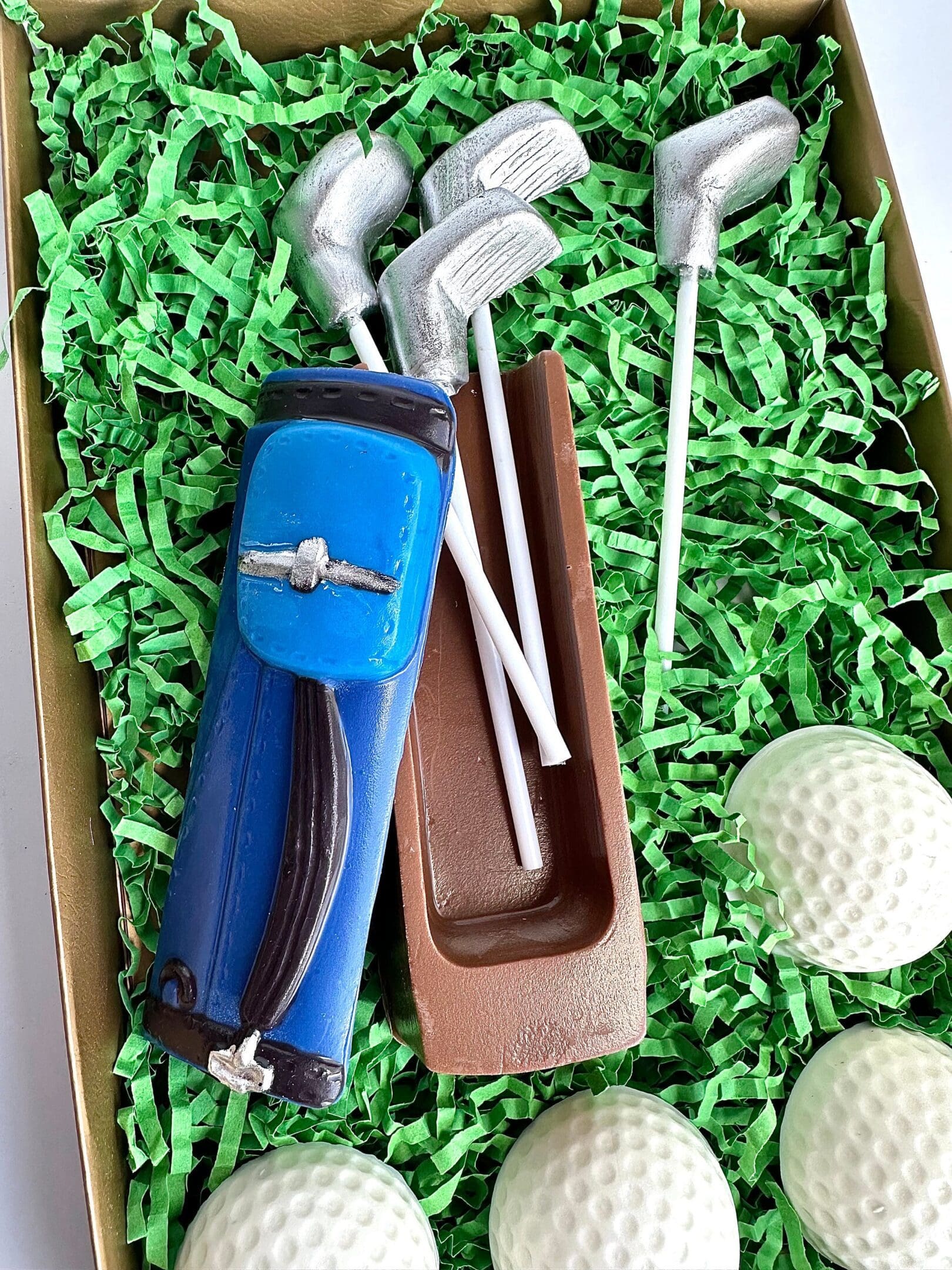 A chocolate gift set of a blue golf bag, clubs, and white golf balls on green shredded paper. Product: Golf Bag With Clubs & Golf Balls Chocolate Gift Set For Dad Birthday Gift/Son Gift/Golf Gift.