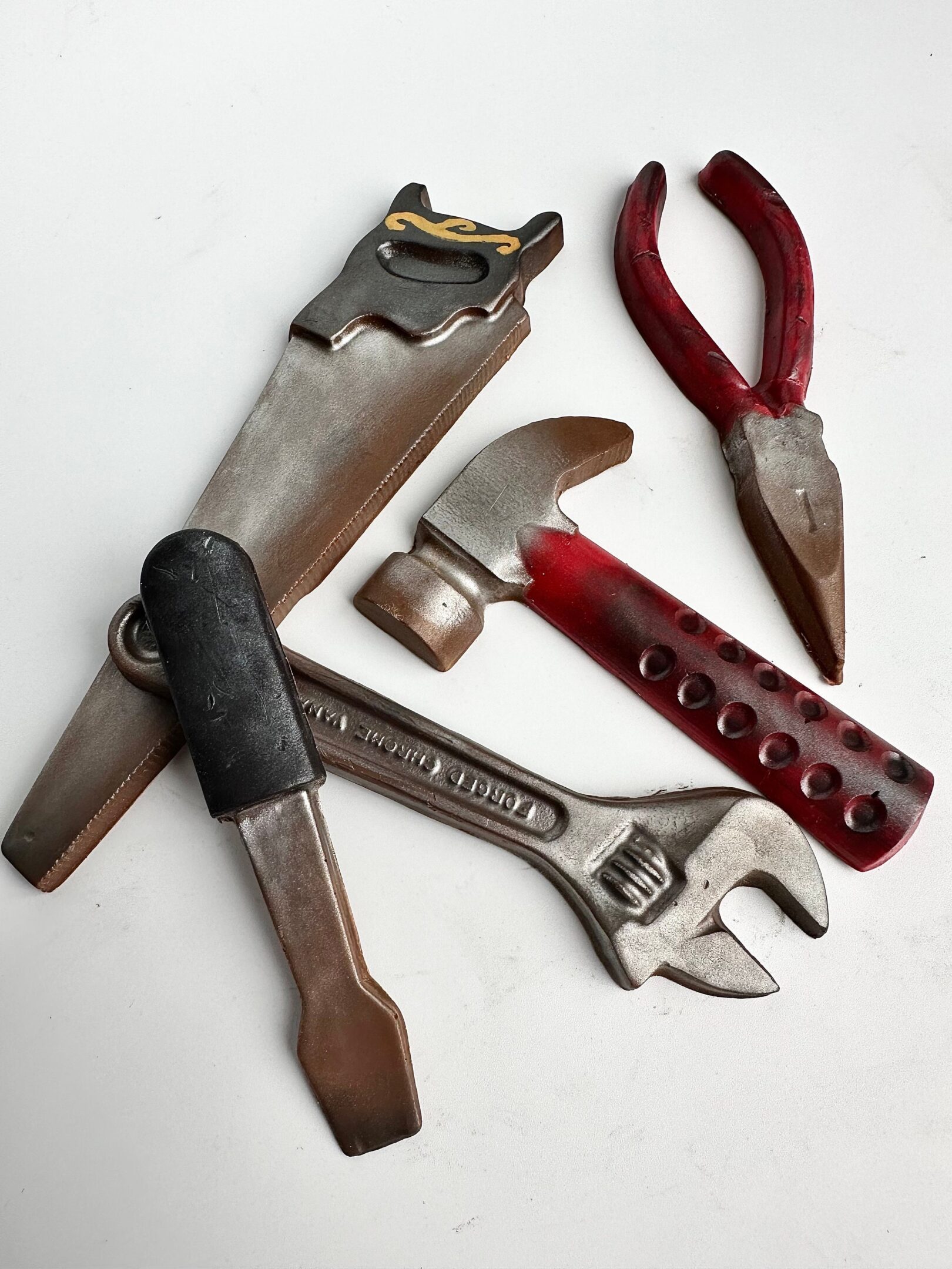 Chocolate candies shaped like a wrench, saw, hammer, and pliers, with a painted metallic appearance, arranged on a white surface.