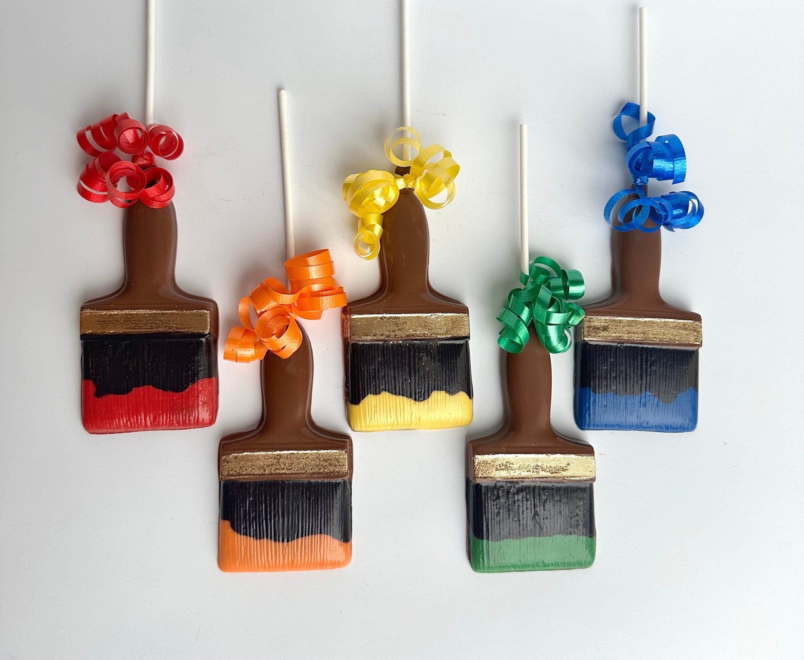 Five chocolate paintbrush lollipops with colored bristles and ribbon decorations in red, orange, yellow, green, and blue against a white background.
