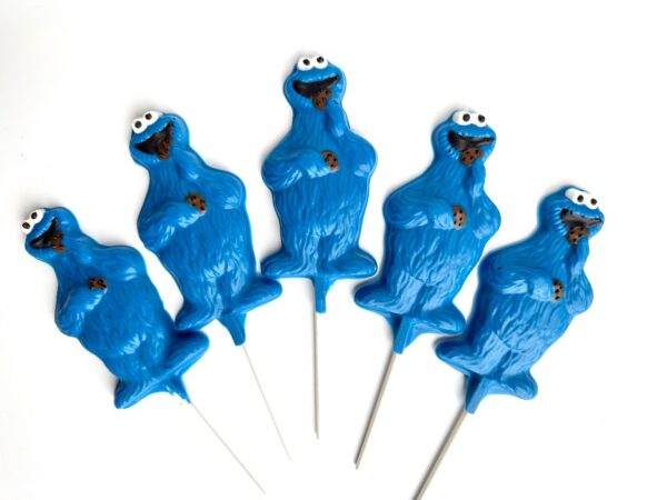 Five chocolate blue monster lollipop favors, resembling cookie monsters holding cookies, are arranged in a row on white sticks against a white background.