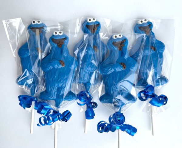 Five blue monster-shaped chocolate lollipops, each holding a cookie, are wrapped in clear plastic with blue ribbon bows at the bottom.