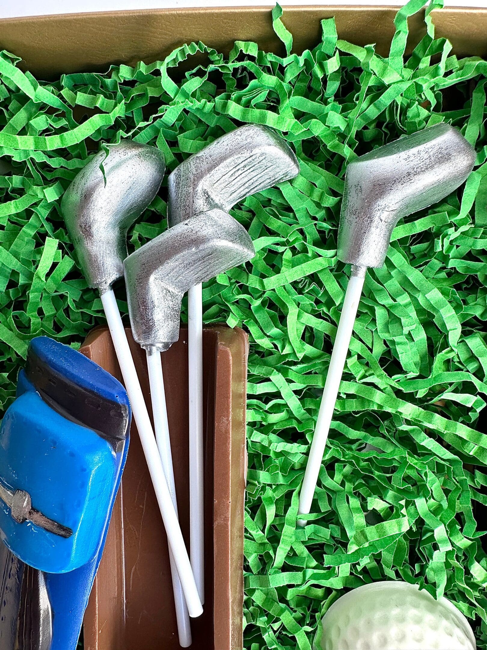 Four silver golf club lollipops, a blue golf bag, a brown block, and a white golf ball on green crinkle paper create the Golf Bag With Clubs & Golf Balls Chocolate Gift Set for Dad Birthday/Son/Golf Gift.