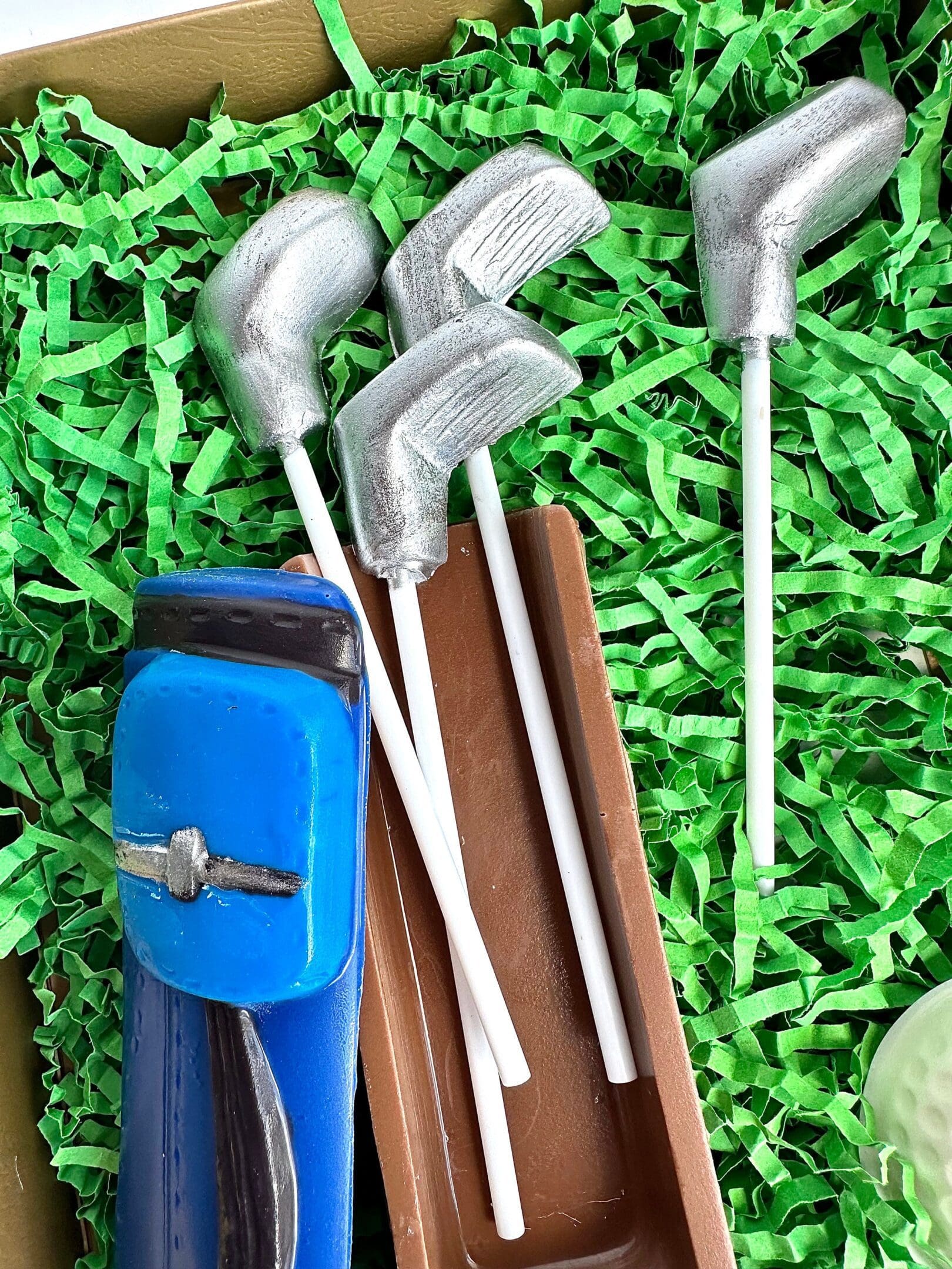 A Golf Bag With Clubs & Golf Balls Chocolate Gift Set featuring four silver golf club cake pops and a blue golf bag on green shredded paper.