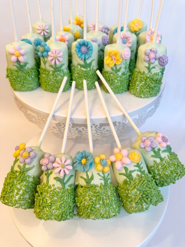 12-flower garden themed chocolate covered double marshmallow pops adorned with edible flowers and green sprinkles on a white stand.