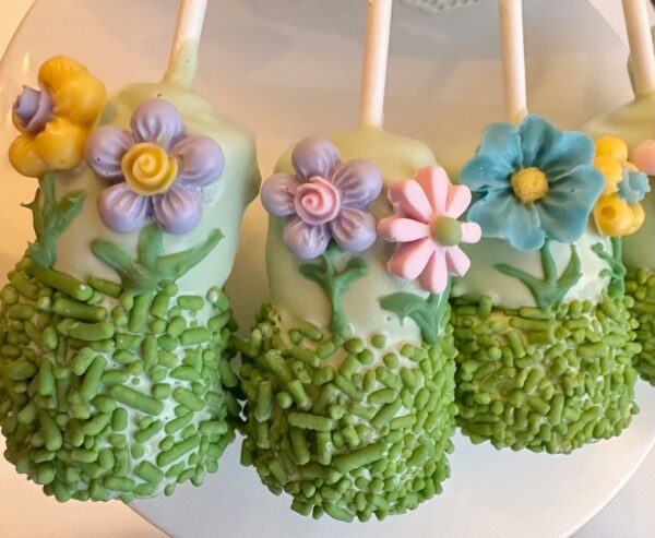 Four garden-themed marshmallow pops, covered in chocolate and decorated with pastel flowers and green sprinkles, arranged on a white surface.