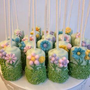 Displayed on a white ornate stand, the 12-Flower Garden Themed Chocolate Covered Double Marshmallow Pops feature white coating, adorned with colorful fondant flowers and green sprinkles.