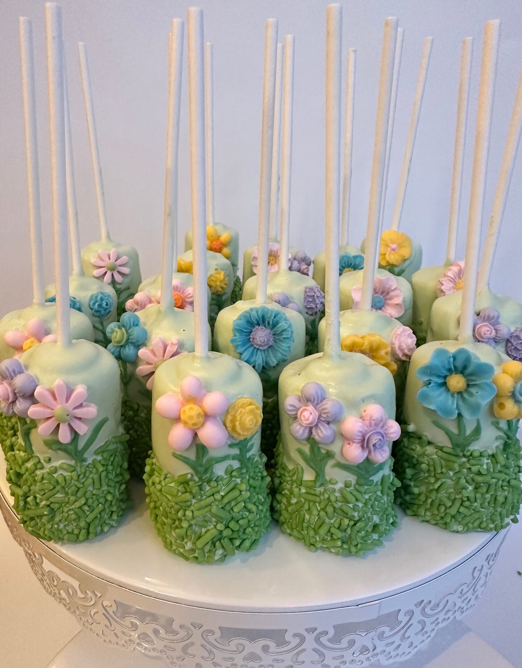 Displayed on a white ornate stand, the 12-Flower Garden Themed Chocolate Covered Double Marshmallow Pops feature white coating, adorned with colorful fondant flowers and green sprinkles.