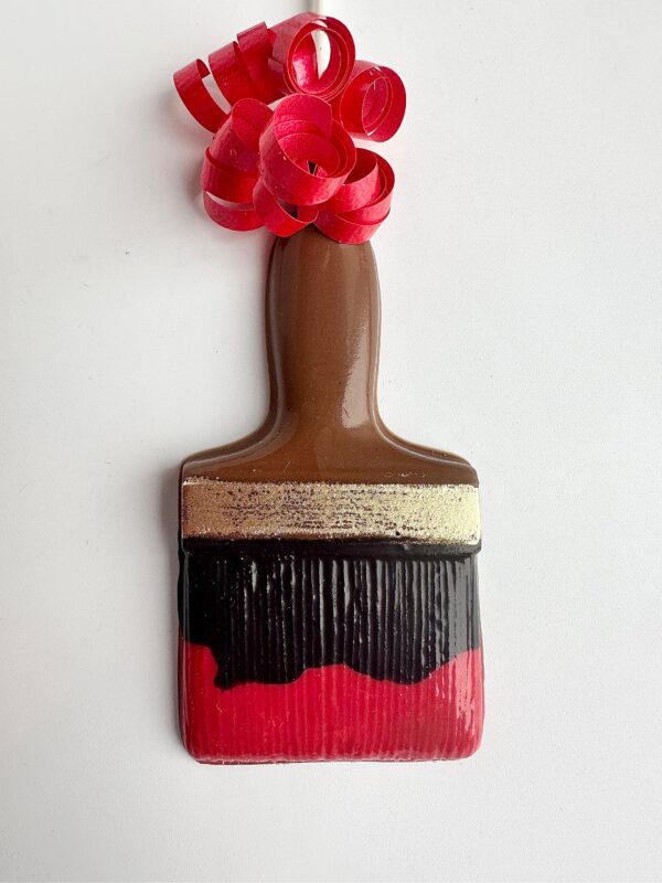 Ceramic paintbrush ornament resembling a chocolate lollipop with red, black, and brown bristles and a wooden-look handle, featuring coiled red ribbon.