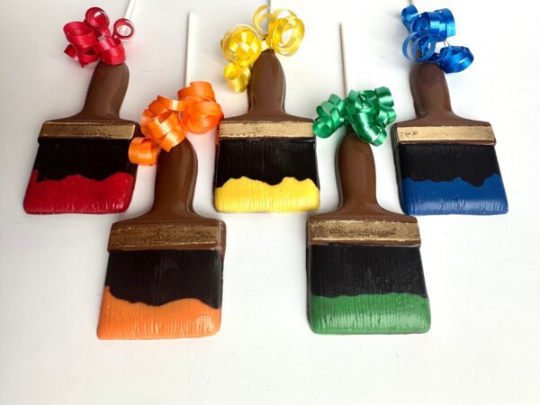 Five chocolate paintbrushes colored red, orange, yellow, green, and blue with matching curled ribbons are arranged on a white surface. They are part of the "12-Paint Brush Chocolate Lollipops for Birthday Party/Party Favors" set.