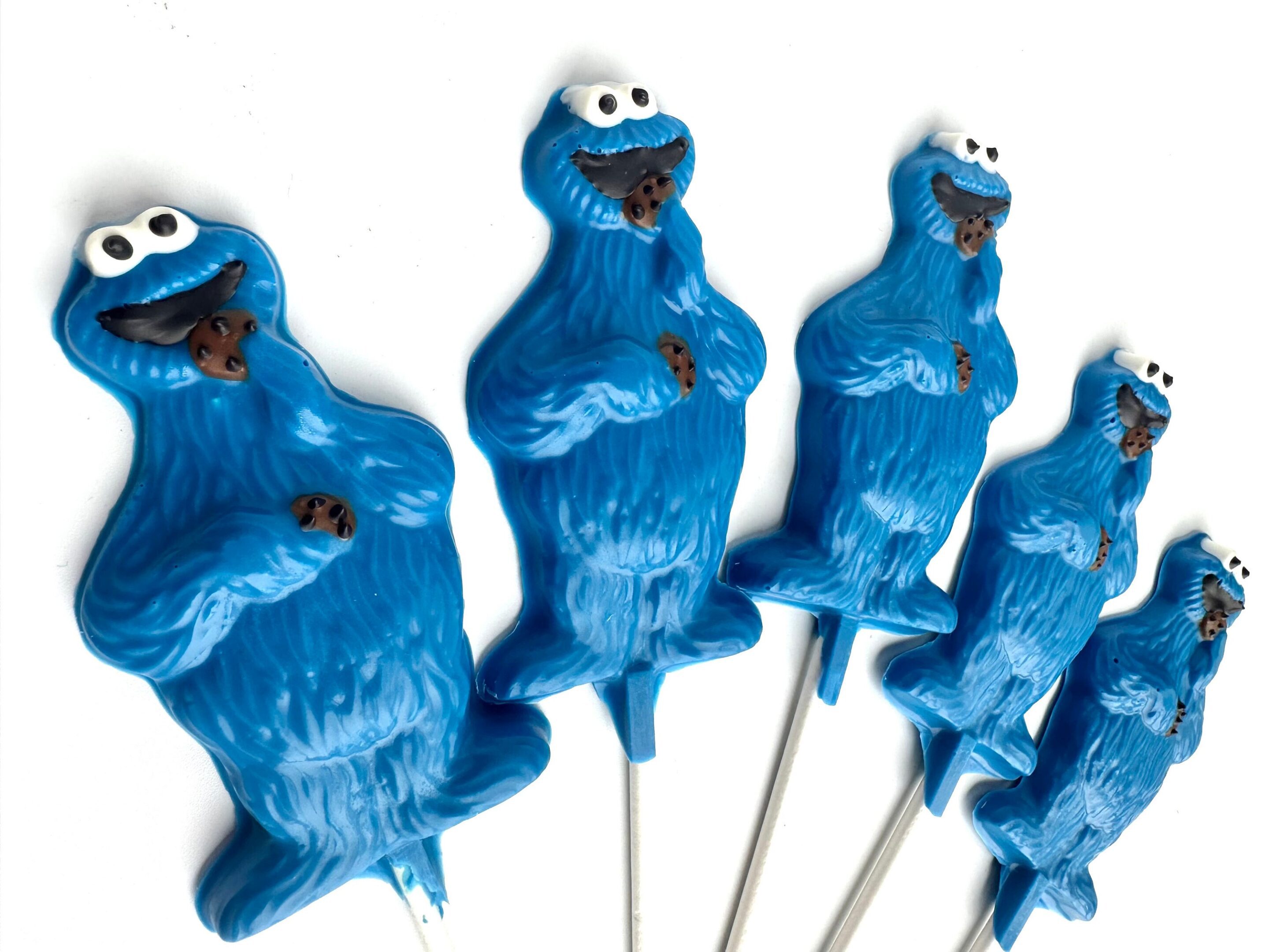 Five blue monster lollipop favors with googly eyes, each holding a brown cookie.