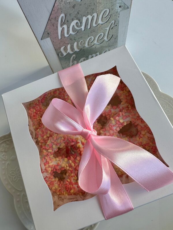 A gift box of 12 strawberry shortcake pretzel twists, decorated with pink sprinkles and tied with a pink ribbon, sits on a white plate. In the background, a "home sweet home" sign is displayed.