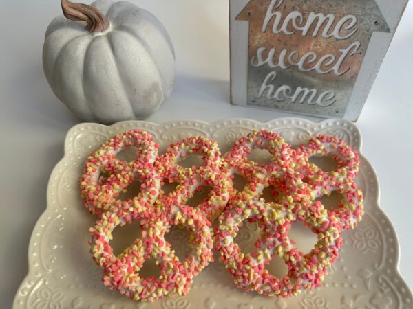 A plate of strawberry shortcake pretzel twists with pink and white sprinkles beside a decorative white pumpkin and a "home sweet home" sign.
