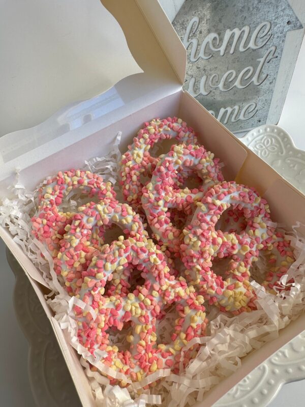 A gift box contains 12 Strawberry Shortcake Pretzel Twists for a birthday or baby shower, topped with white chocolate and pink and yellow sprinkles, nestled on shredded paper. A "home sweet home" sign is nearby.