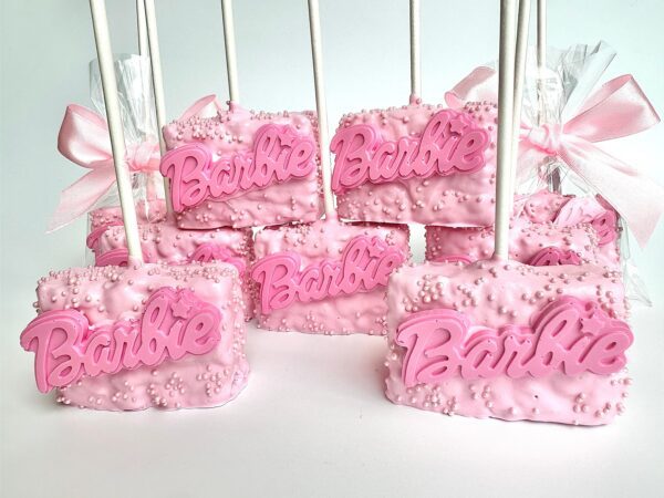 Pink dessert pops are chocolate-covered Rice Krispie treats themed with "Barbi" text and decorative sprinkles, each carefully wrapped individually with a white stick and adorned with a pink ribbon.