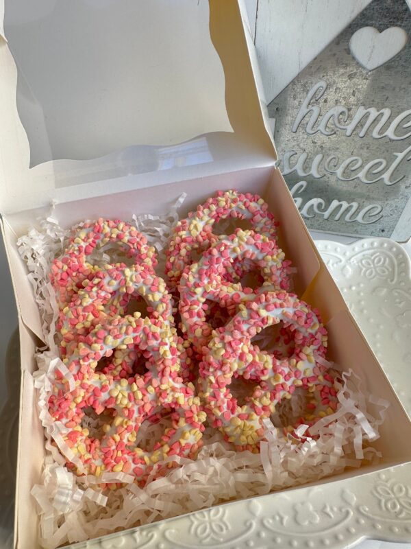 A pastel box contains 12 strawberry shortcake pretzel twists adorned with pink and yellow sprinkles, neatly arranged on shredded white paper. A background sign reads "home sweet home." Perfect for a birthday, baby shower, or welcoming a baby girl.