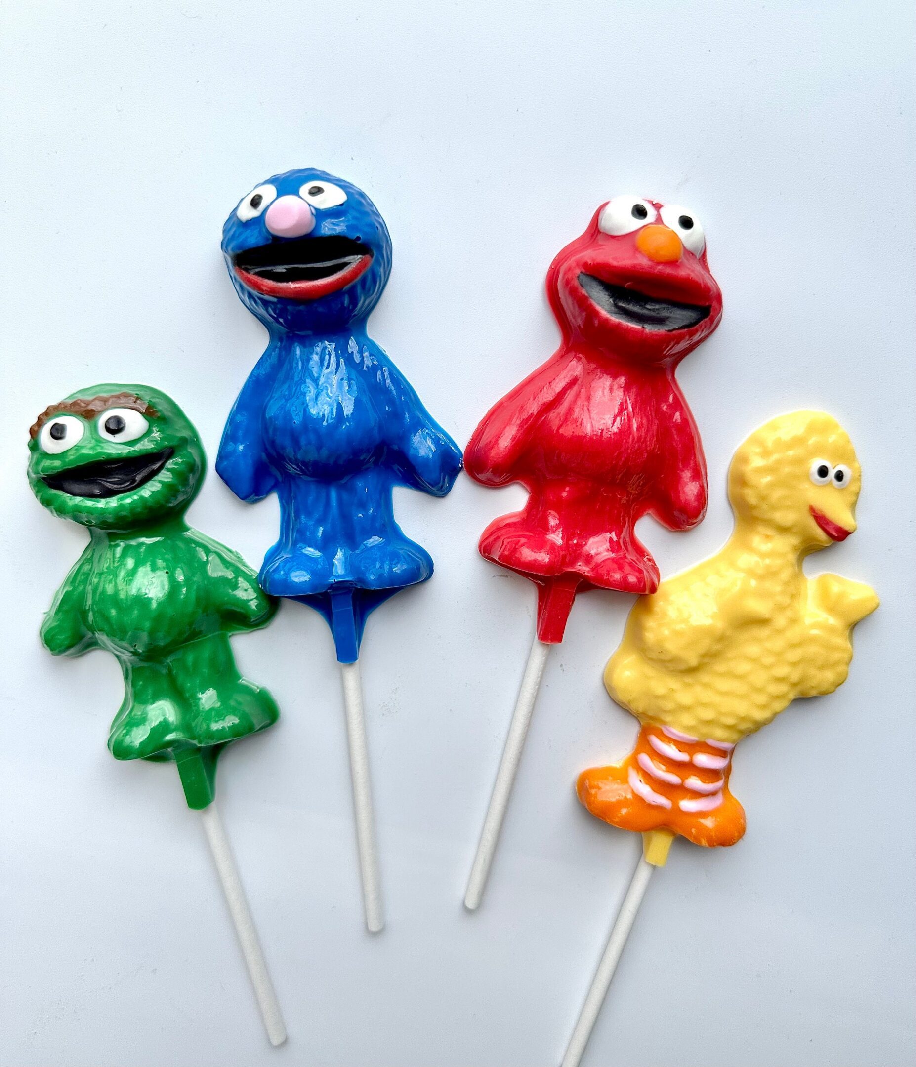 Four Sesame character lollipops in blue, red, green, and yellow on white sticks are displayed on a white background. Part of the 12-Assorted Sesame Character Lollipop Favors for Birthday Party/Party Favor/Children’s Party set.
