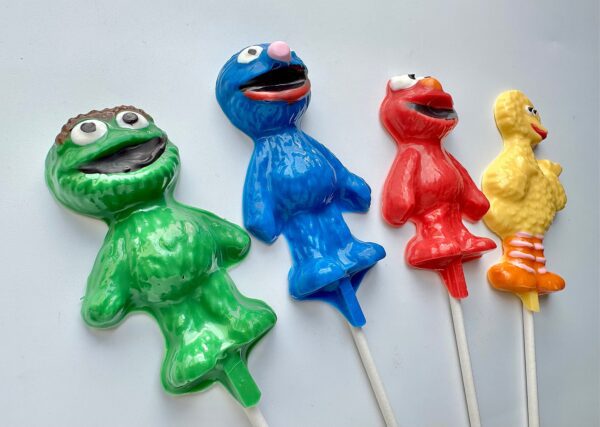 Four colorful Sesame character-shaped lollipops—green, blue, red, and yellow—are lined up with unique facial features and poses against a plain white background.