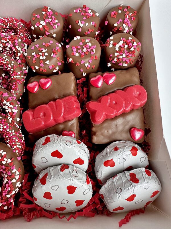 A Milk Chocolate Treat Gift Box features chocolate-covered treats adorned with pink and red heart sprinkles, some inscribed with "LOVE" and "XOXO" in red icing. Perfect for anniversaries, Valentine’s Day, or birthdays.