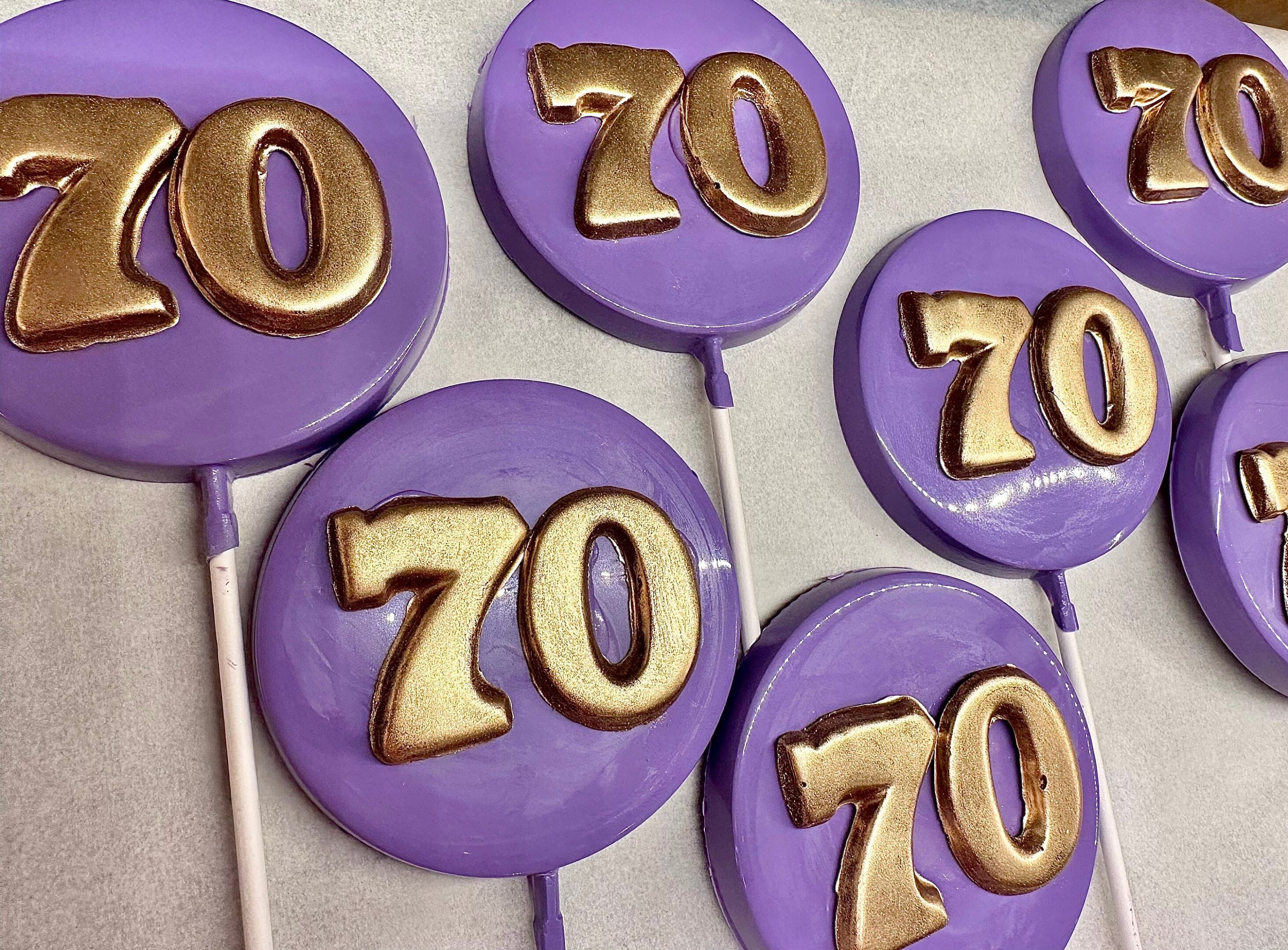 Purple lollipops with gold "70" decoration from the "12-Purple 70th Birthday/Anniversary Chocolate Lollipop/Party Favors/70th Birthday Party" set are laid on a white surface.