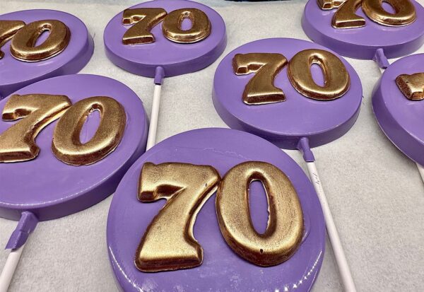 Purple chocolate lollipops feature gold "70" numbers, designed for a 70th birthday or anniversary party, displayed on a white surface.