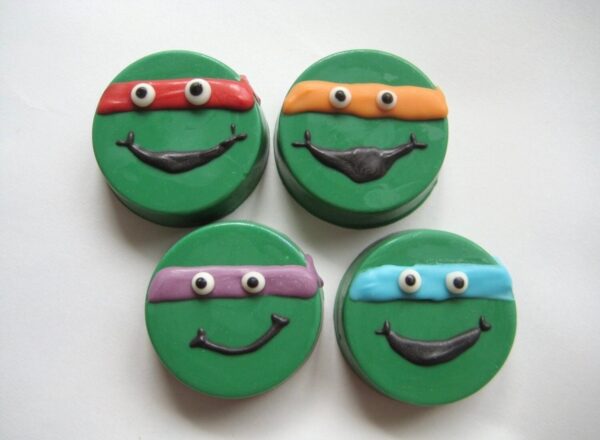 Four green smiley face cookies shaped like ninja turtles with chocolate-covered Oreos, each wearing a red, orange, purple, or blue mask for a superhero-themed birthday party on a white background.