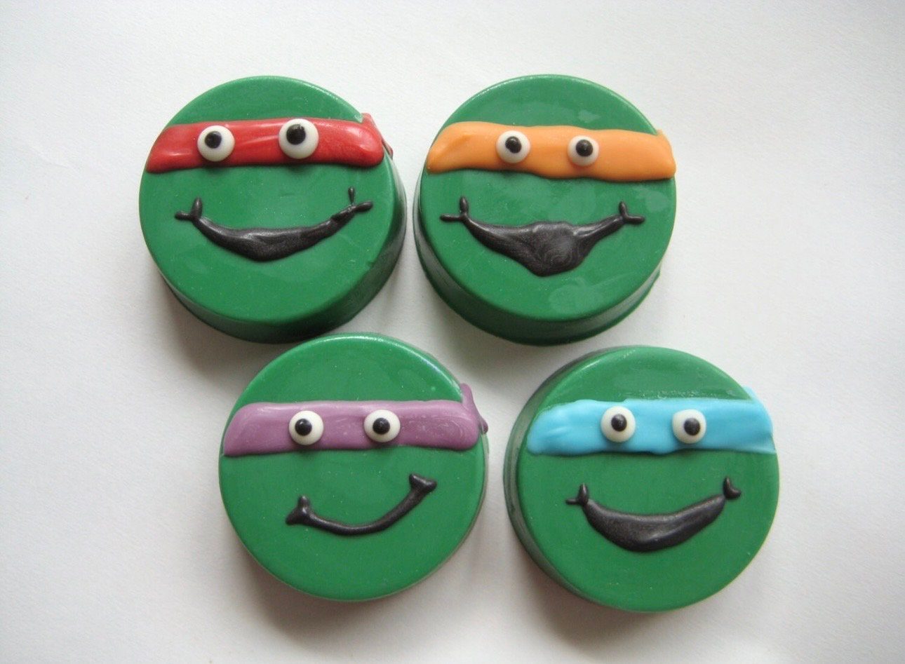 Four green smiley face cookies shaped like ninja turtles with chocolate-covered Oreos, each wearing a red, orange, purple, or blue mask for a superhero-themed birthday party on a white background.