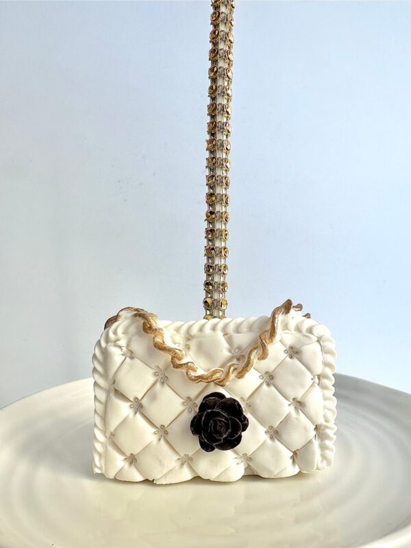 A small white quilted cake shaped like a designer purse from the "12-Designer LOGO Purses" collection, featuring a gold chain handle and black flower decoration, displayed on a white plate.