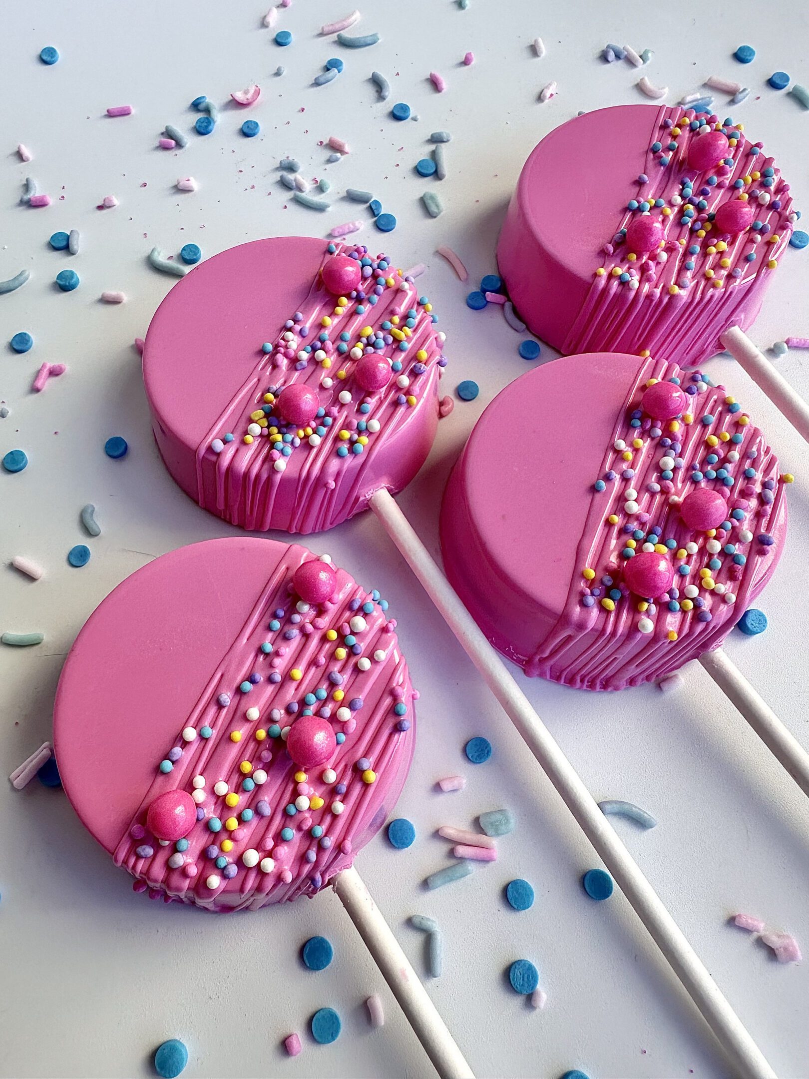 Four hot pink Oreo lollipops with sprinkles and drizzle on a white surface, surrounded by scattered sprinkles.
