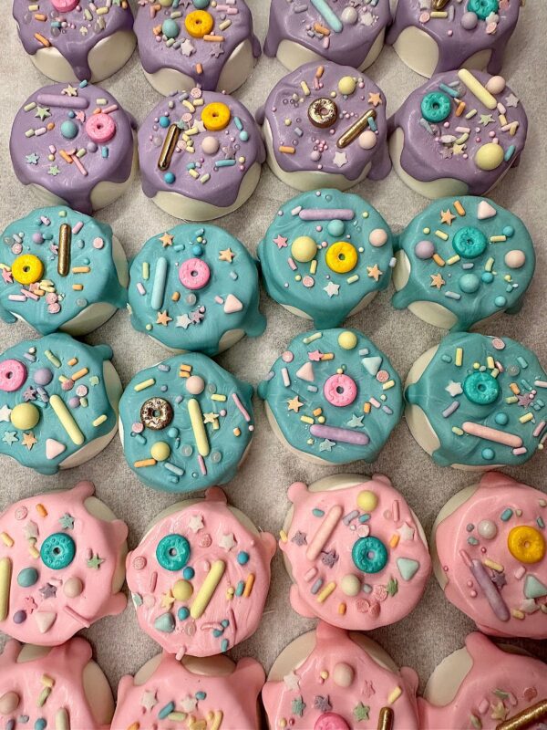 Drippy chocolate-covered Oreos, decorated with pastel blue, purple, and pink icing topped with colorful sprinkles, stars, and candy shapes are arranged in rows on a tray for a birthday party or baby shower.