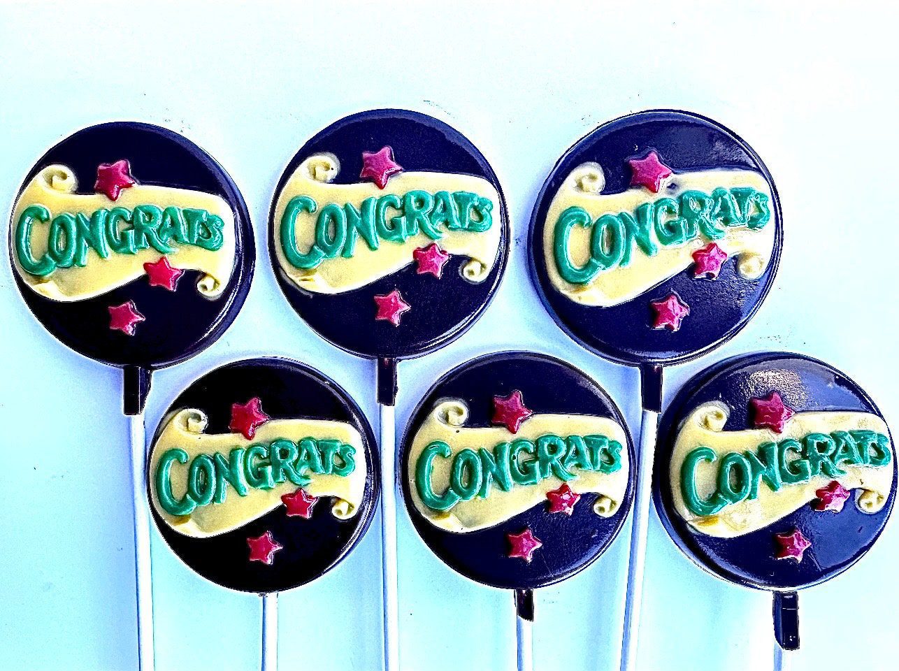 Six chocolate lollipops with "Congrats" designs in green and yellow text on black, featuring red stars. (Product: 12-Congratulations Chocolate Lollipops).