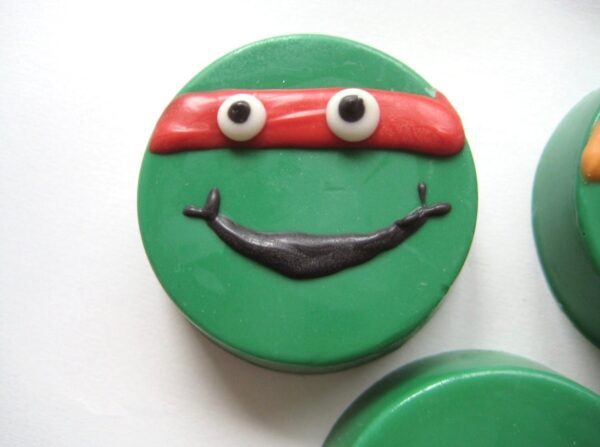 A green circular item featuring a smiling face with googly eyes, a red band, and a black smile on white background is the 12-Ninja Green Turtles Chocolate Covered Oreos for Birthday Party/Party Favors/Super Hero Theme.