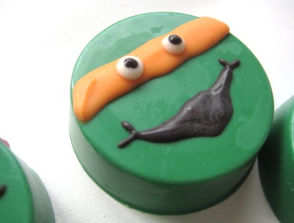 Green round cookie decorated with orange band, white eyes, and a black curved mouth, resembling a cartoon face from the '12-Ninja Green Turtles Chocolate Covered Oreos for Birthday Party'.