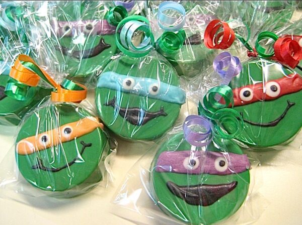 Green cookies designed as cartoon faces, each wrapped in clear plastic with colorful ribbon bows. Product: 12-Ninja Green Turtles Chocolate Covered Oreos for birthday or superhero-themed party favors.