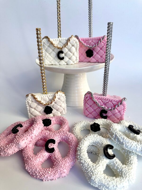 A stand displays decorative cake pops shaped like purses, featuring a "C" emblem and covered in pink and white icing with chains and pretzels, from the "12-Designer LOGO Purses" collection perfect for events.
