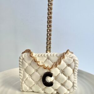 A white, quilted cake shaped like a designer purse with a gold chain handle and black "C" emblem is presented on a plate.