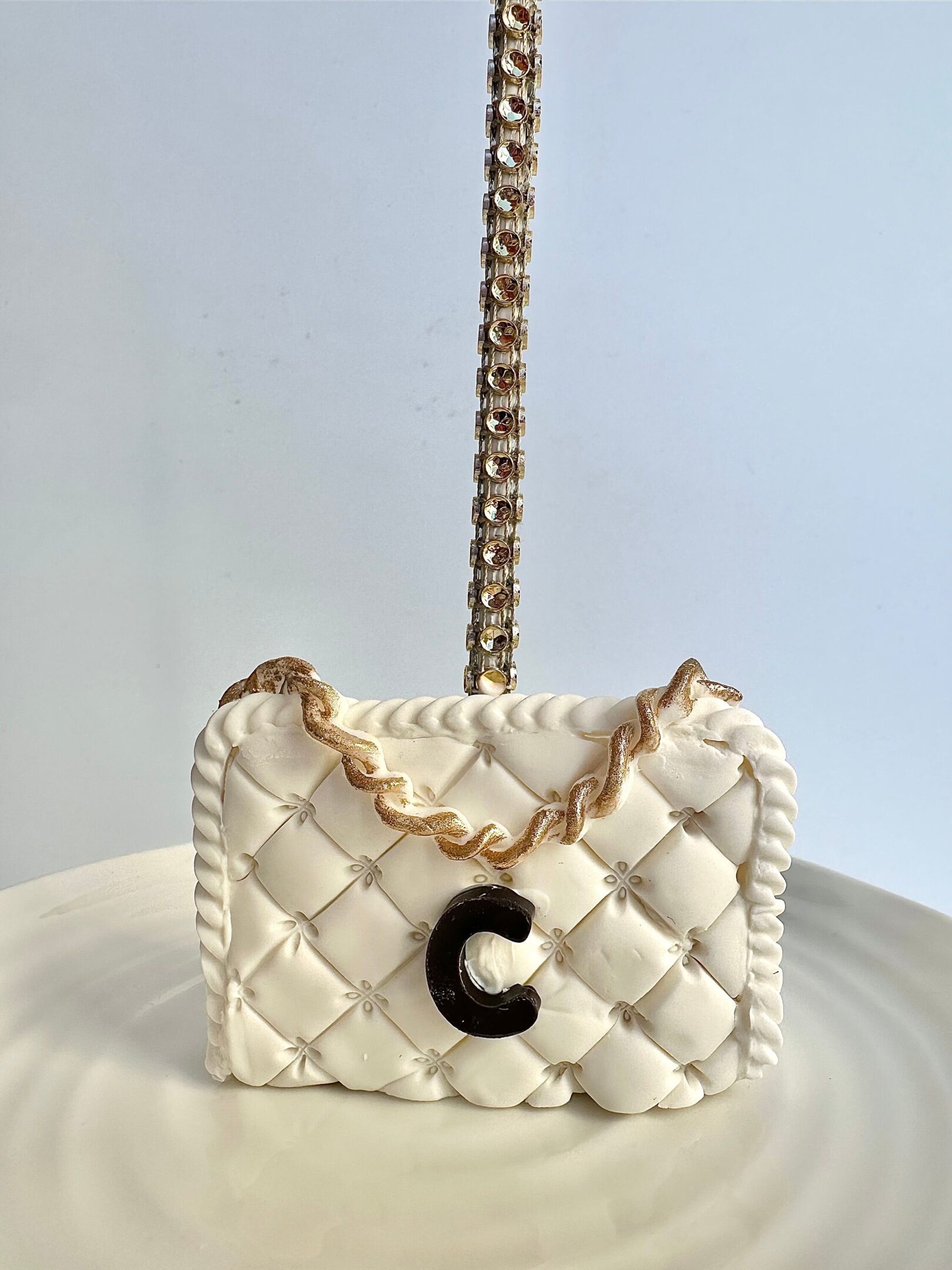 A white, quilted cake shaped like a designer purse with a gold chain handle and black "C" emblem is presented on a plate.