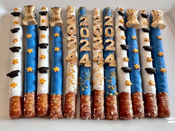 Pretzel rods dipped in blue and white frosting with gold stars and bells featuring "2024" for graduation, named 12-Graduation 2024 Gold Diploma Chocolate Covered Pretzel Rods For Graduation Party/Graduation Favors.