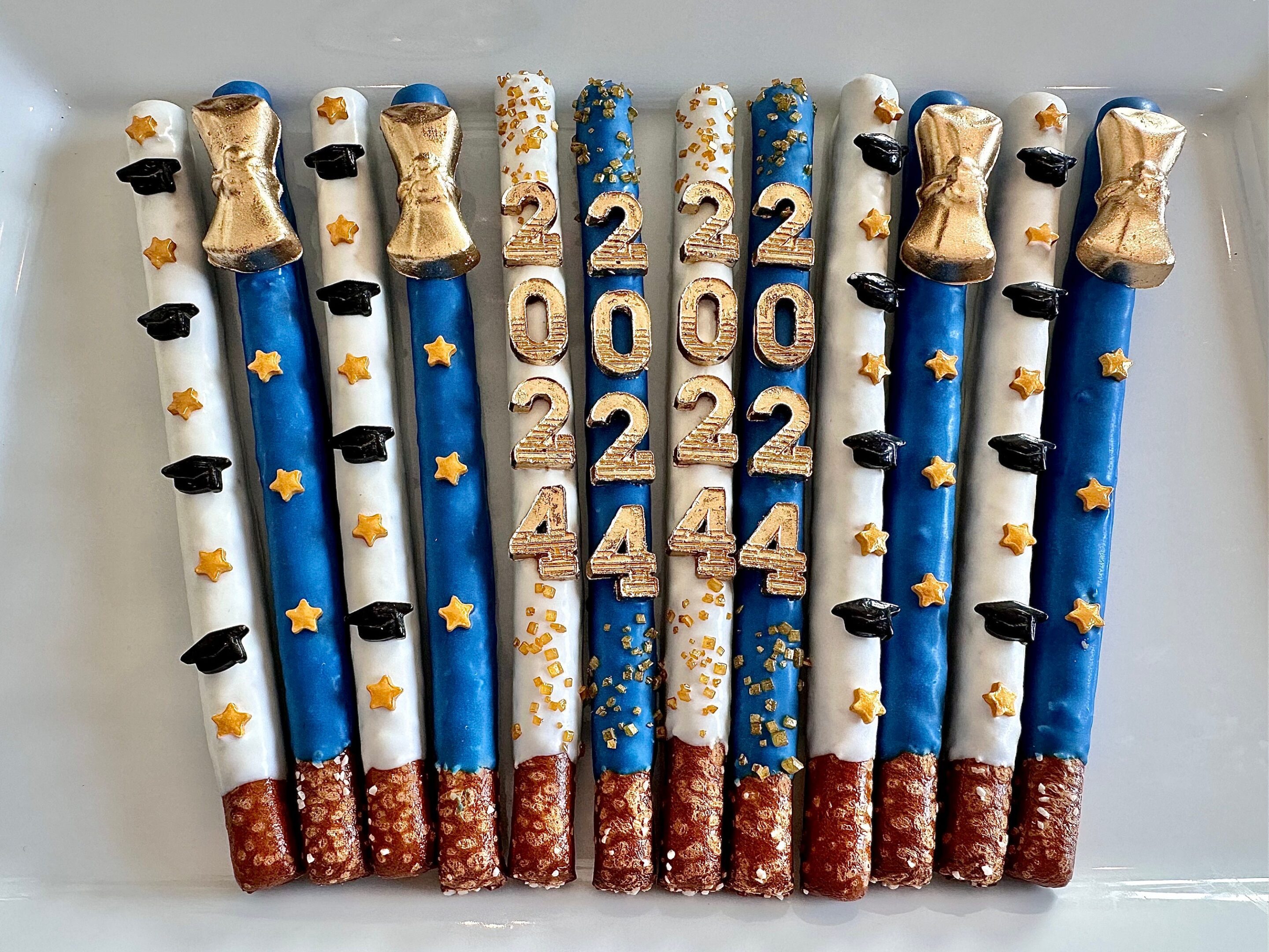 The 12-Graduation 2024 Gold Diploma Chocolate Covered Pretzel Rods feature blue, white, and red coatings with golden stars, graduation caps, and the numbers 2022 and 2024 in gold for your graduation party or favors.