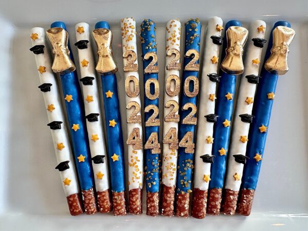 Blue, white, and red iced pretzel rods, decorated with stars, glitter numbers, and gold hourglass accents—12-Graduation 2024 Gold Diploma Chocolate Covered Pretzel Rods for your graduation party—elegantly displayed on a tray.