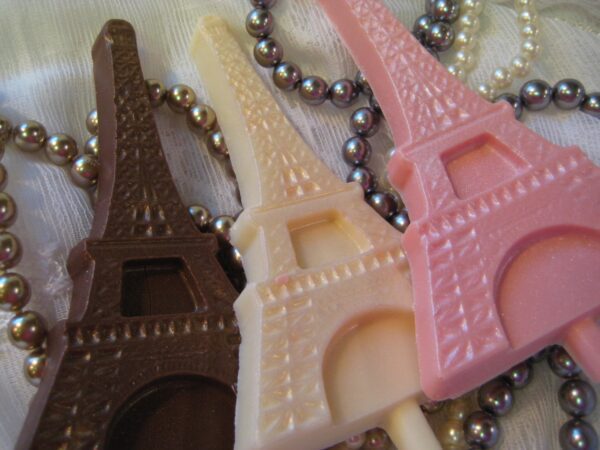 Three Eiffel Tower-shaped chocolates in dark brown, cream, and pink from the 12-Chocolate Eiffel Tower Lollipop Favors collection, surrounded by pearl-like beads on a white fabric background.