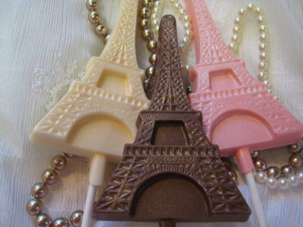 12-Chocolate Eiffel Tower Lollipop Favors in brown, cream, and pink are displayed on a decorative fabric background adorned with pearl-like beads.
