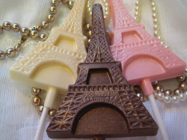 Three Chocolate Eiffel Tower Lollipops in white, dark, and pink from the 12-Chocolate Eiffel Tower Lollipop Favors set are displayed with pearl necklaces on a textured surface.