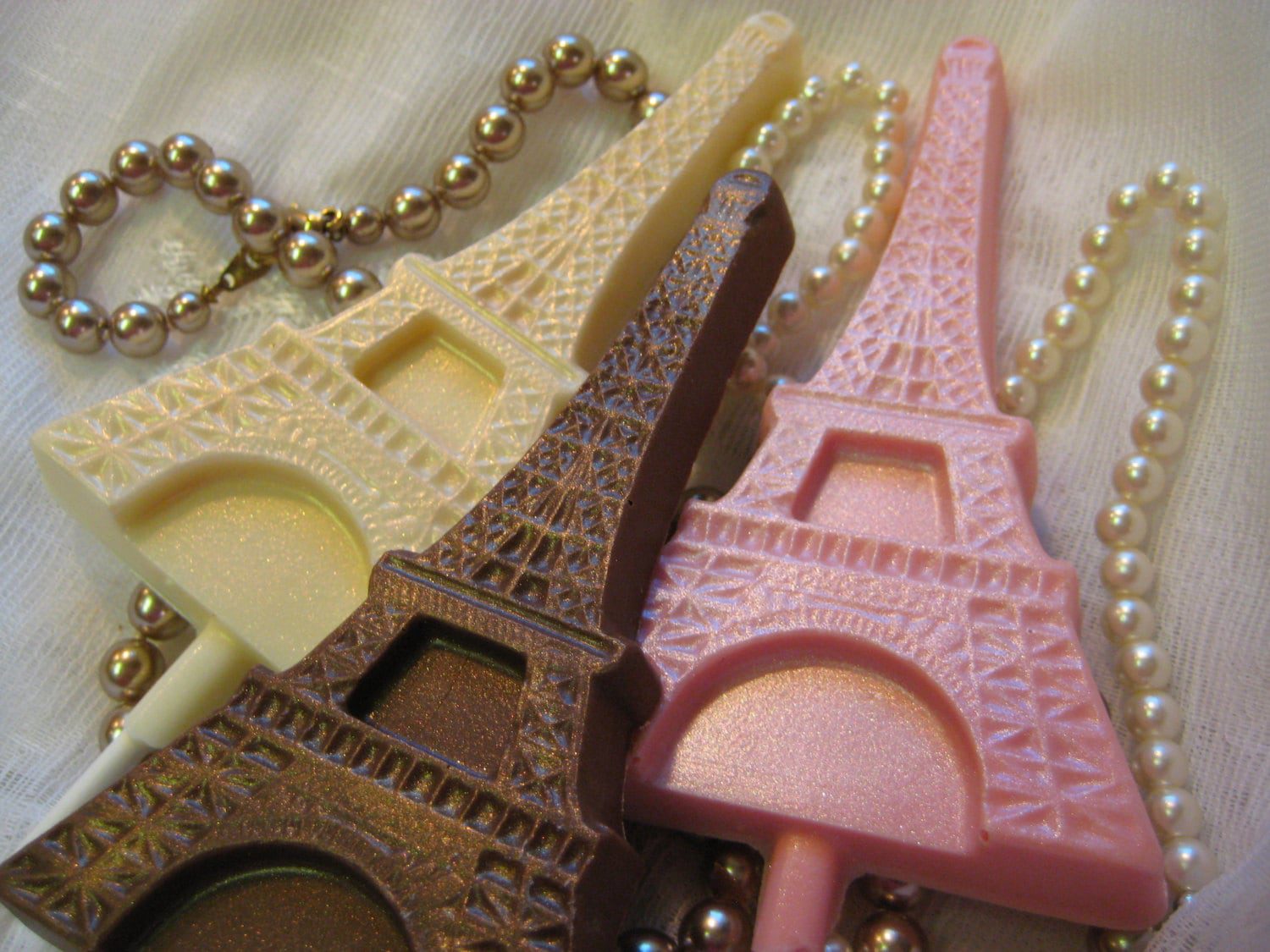 White, brown, and pink 12-Chocolate Eiffel Tower Lollipop Favors are displayed on a white fabric adorned with strings of pearls.