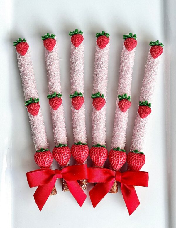 Six pink candy-coated pretzel rods with strawberry tops and red bows, vertically arranged on a white background.