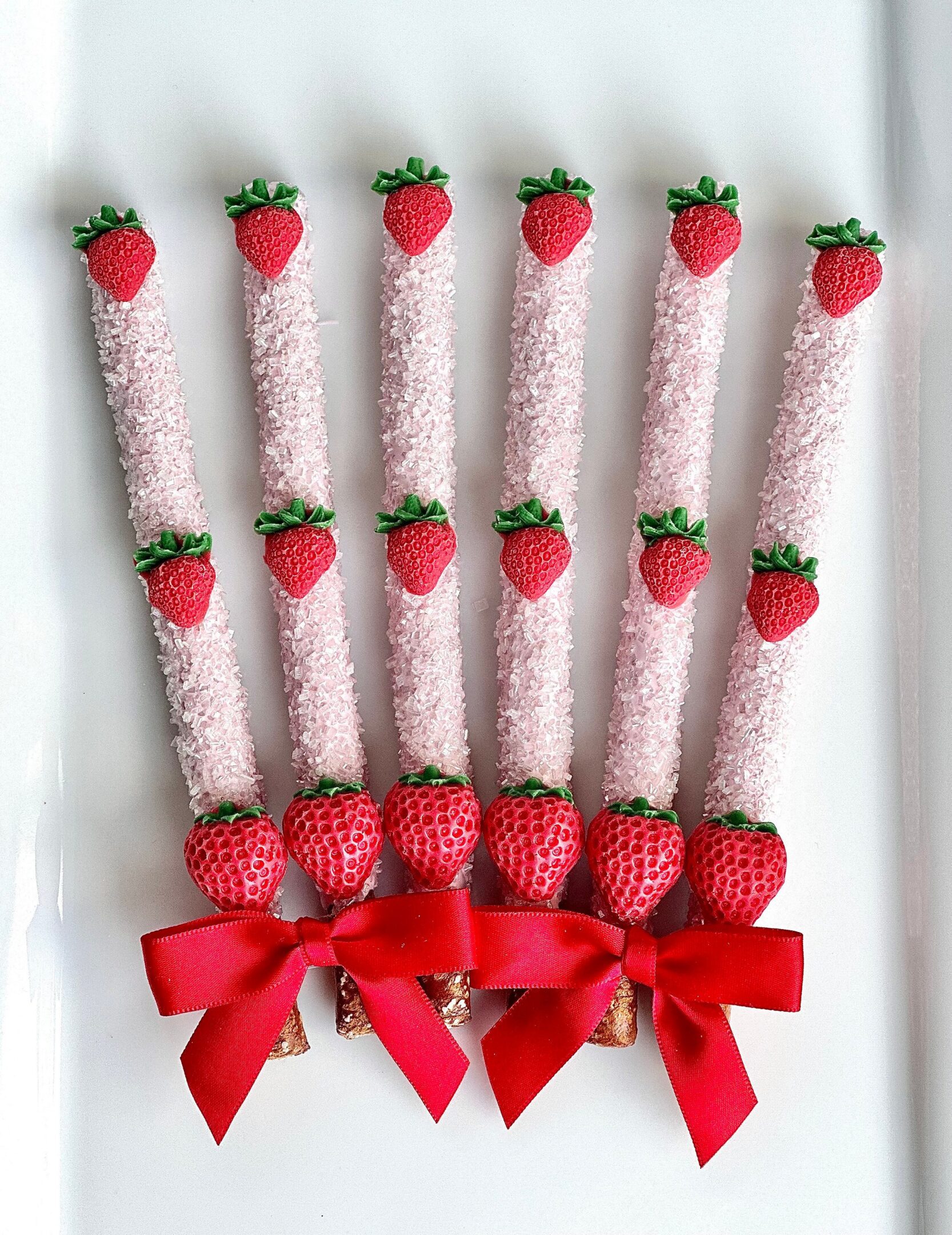 Six pink candy-coated pretzel rods with strawberry tops and red bows, vertically arranged on a white background.
